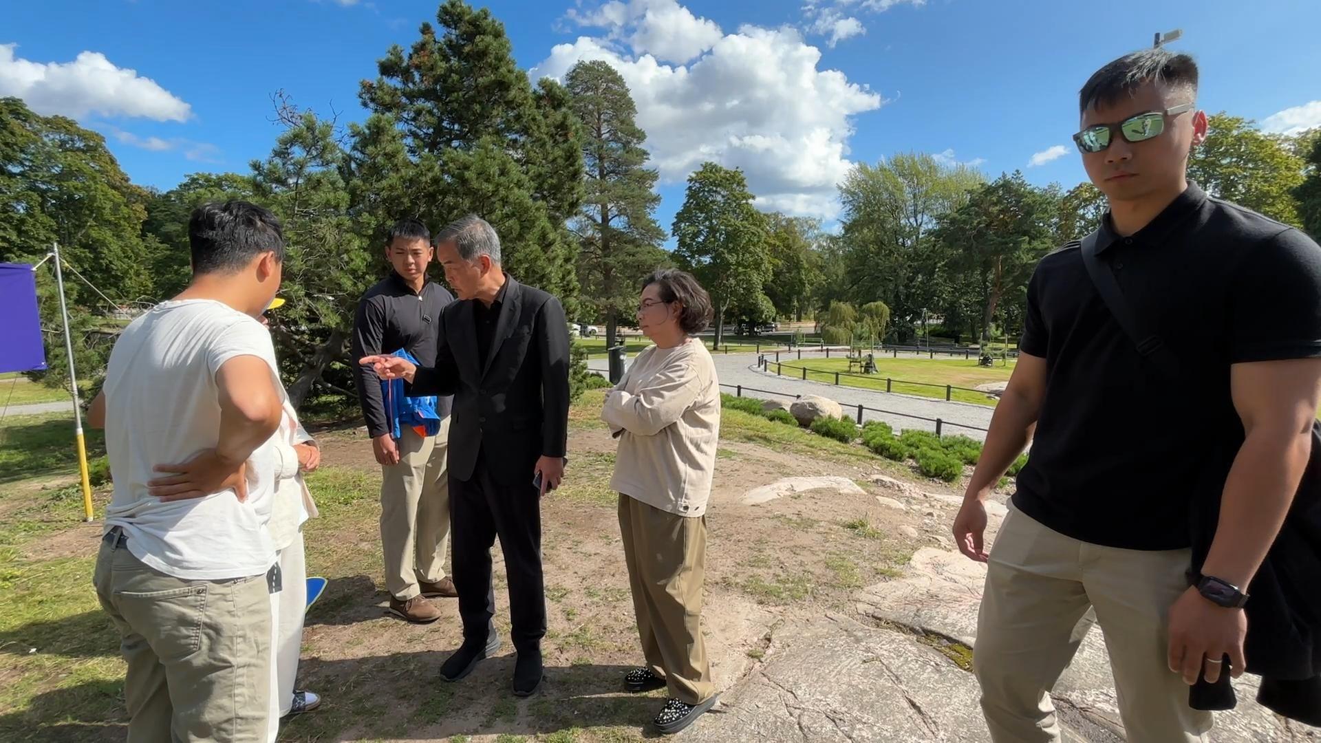 芬蘭赫爾辛基西貝柳斯公園內，身穿黑色西裝的梁振英（中左）指罵兩名身穿白色上衣的法輪功學員（左），身穿米白色上衣的夫人梁唐青儀（中右）在旁注視，兩名身穿黑色上衣的疑似保鑣站在梁振英夫婦左右兩側（法輪大法信息中心發放影片截屏13/8/2025）