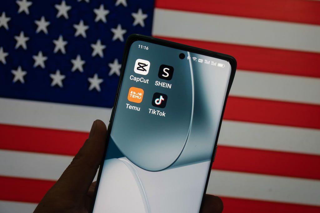 Una mano sostiene un teléfono donde se ven los logos de Temu y Shein y en el fondo la bandera de EE.UU.