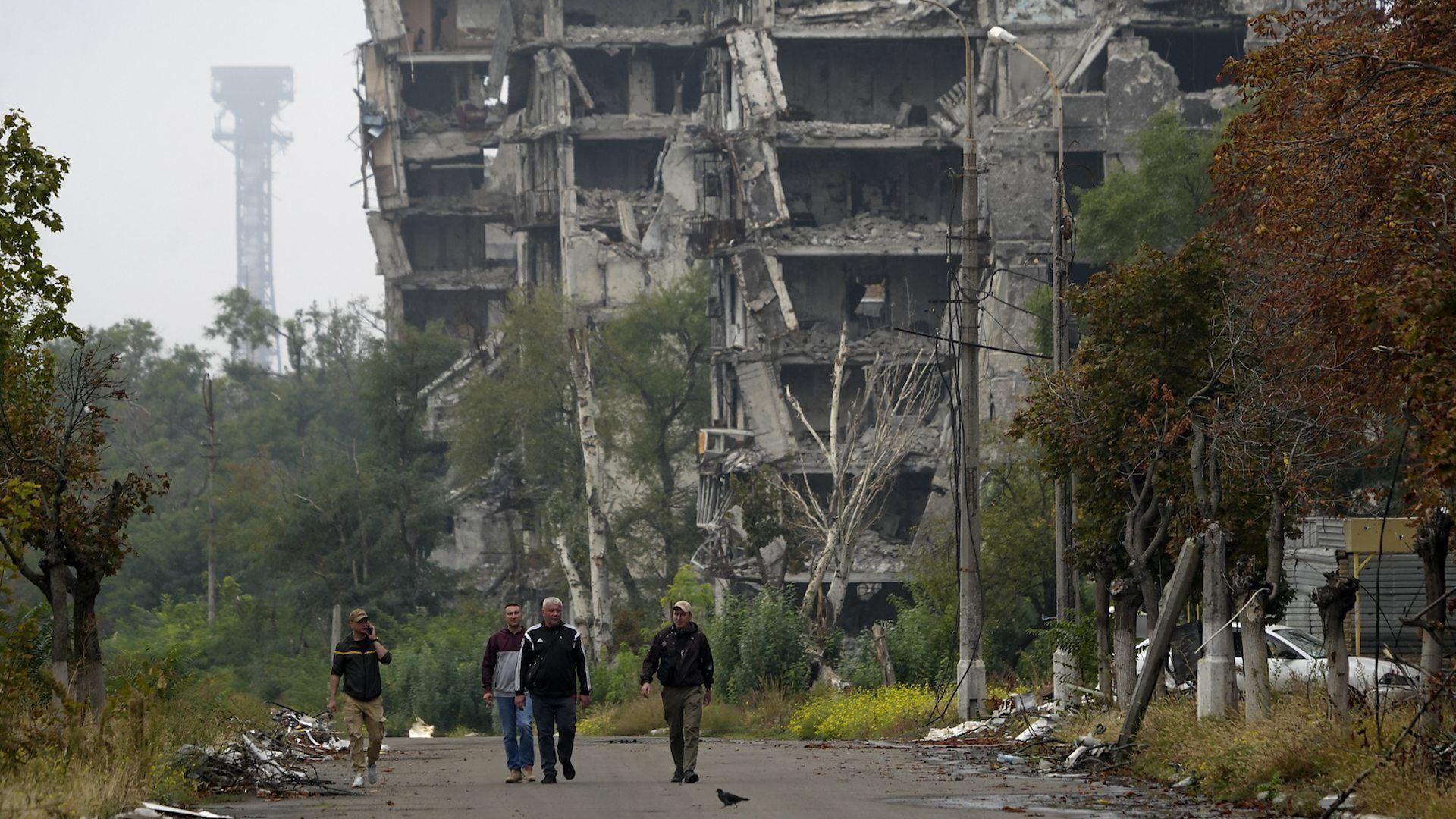 Una desolada calle de Mariupol, en la que un edificio en ruinas por los bombardeos de la guerra y una lejana torre de metal que se erige sobre un verde bosque le sirven de fondo lúgubre a la imagen de cuatro hombres que caminan por la calle, en medio de los escombros y la naturaleza descuidada.