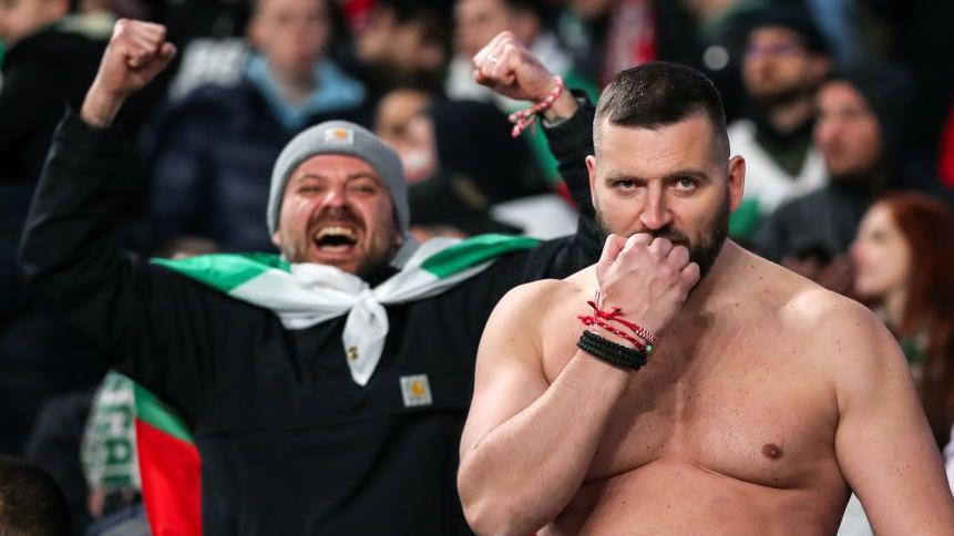 مشجعان لكرة القدم في مدرج على اليمين شاب بلحية دون قميص يرفع قبضته إلى فمه، على اليسار شاب يضع علم فريق حول رقبته، برتدي قبعة ويرفه يديه محتفلاً