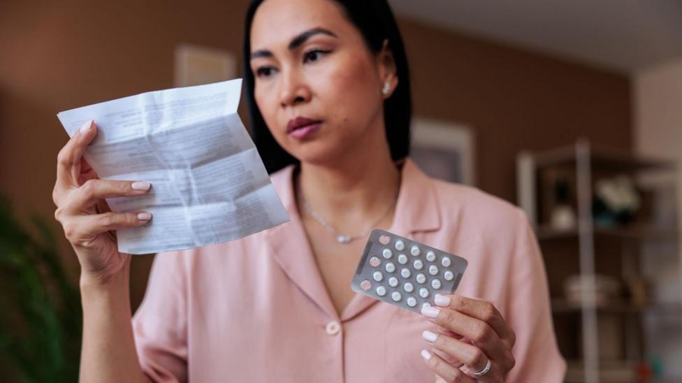 Una mujer con una camisa rosada sostiene en una mano un blister de pastillas mientras lee el prospecto de ese medicamento que sostiene con su otra mano