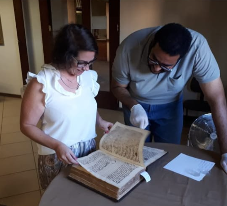 Pesquisadores analisam um manuscrito antigo em uma mesa de trabalho. Ela folheia as páginas enquanto ele observa atentamente, usando luvas de proteção para manusear o documento histórico. 