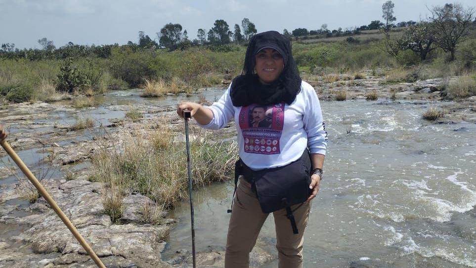 Yadira González de pie cerca de un riachuelo, con una camisa que tiene la imagen de su hermano. Le sonríe a la cámara y sostiene una especie de bastón como los que se usan para caminar en montañas.