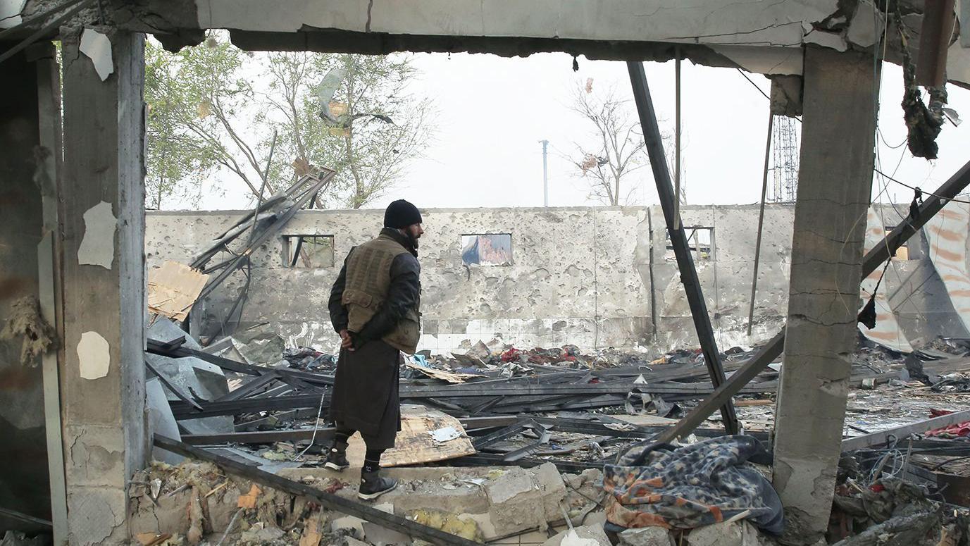 A man stands in the burnt wreckage of the rehabilitation facility. His back is ot the camera. The walls are blackened. Sky can be seen through the broken roof, there is rubble on the floor.