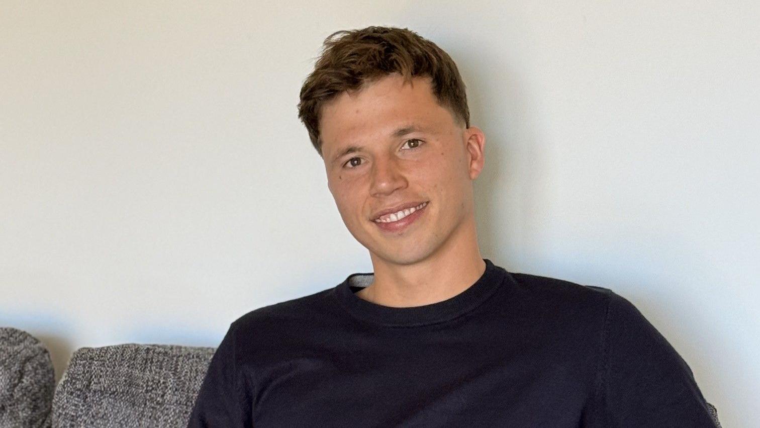 Freddie Woodward: un joven de pelo corto y oscuro está sentado en un sofá gris, sonriendo a la cámara. Tiene el pelo corto castaño y lleva un jersey azul marino.