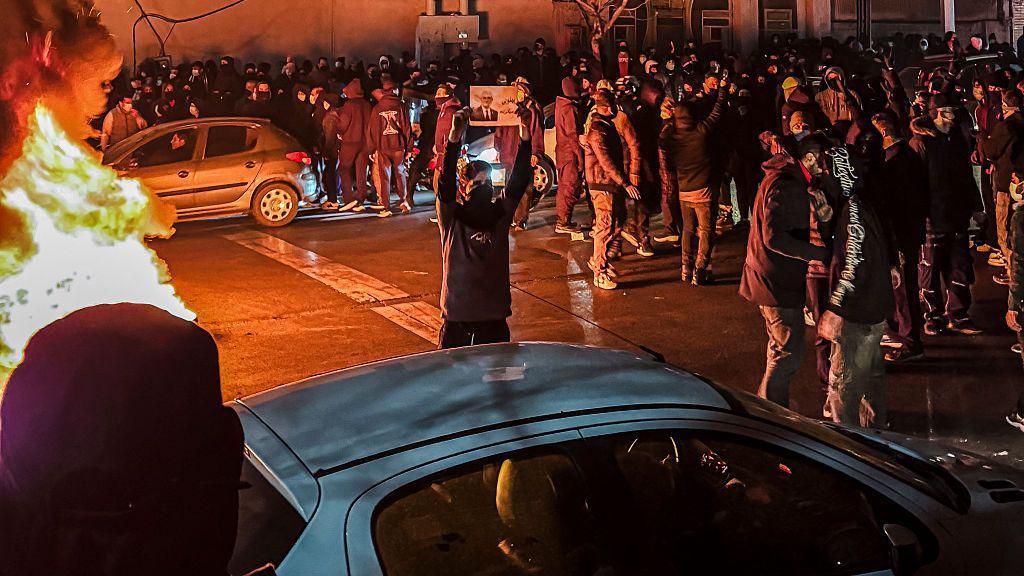 Manifestantes se re&uacute;nem nas ruas de Teer&atilde;, no Ir&atilde;. No centro da imagem, um manifestante mostra uma fotografia. No lado esquerdo, o fogo queima soltando fuma&ccedil;a.