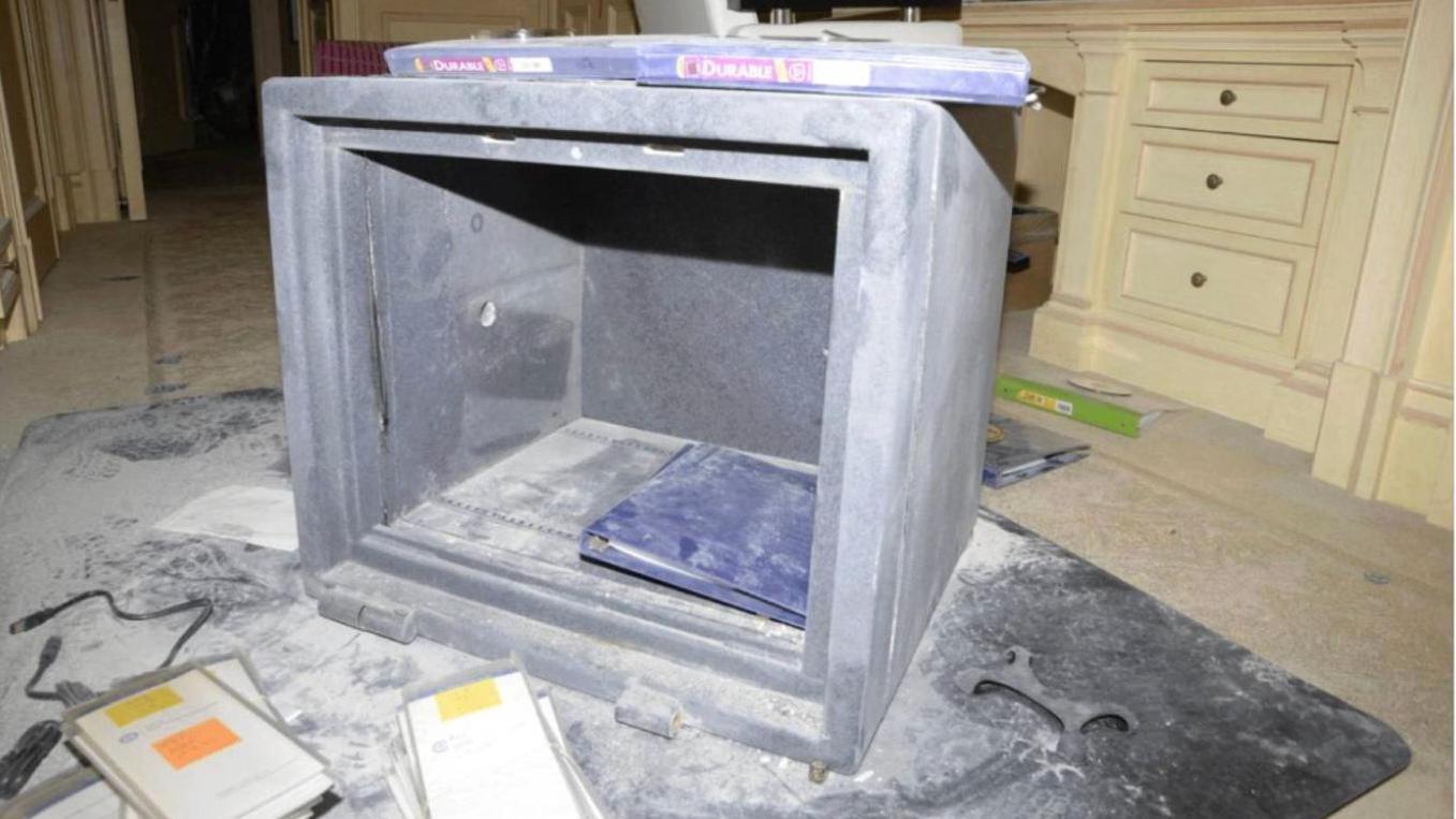 Una caja fuerte con la puerta rota sobre un suelo alfombrado, con polvo esparcido por todas partes y lo que parecen ser muebles de dormitorio de color crema con cajones incorporados en el fondo.