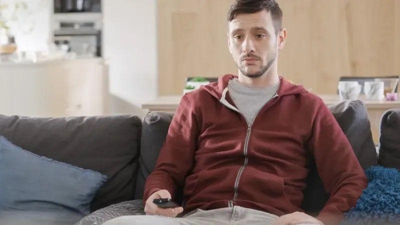 Um homem olhando para a TV com rosto de tédio e segurando um controle remoto