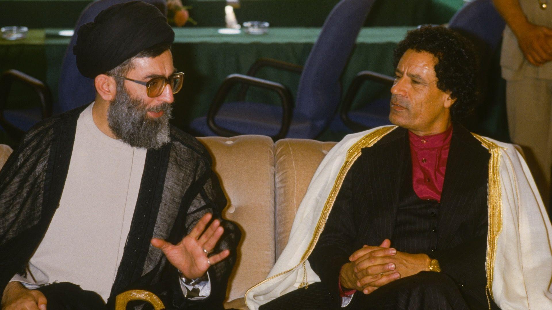 Gaddafi, sitting to the right of the image, looks at Khamenei as the Iranian leader talks. Gaddafi is holding his hands together on his lap. He is wearing a white robe with golden embroidery on the sides, a black jacket and dark red shirt with no collar, buttoned to the top. Khamenei is holding his hand up as he speaks. He is wearing black clothing on his head, a grey and black jacket and cream shirt. The pair are sitting next to each other on a sofa, with desk chairs and a meeting table behind them.