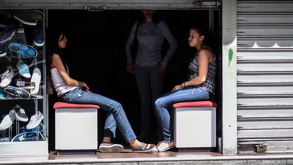 Funcion&aacute;rias de uma sapataria sentadas na entrada de uma loja &agrave;s escuras, durante um corte de eletricidade em Caracas