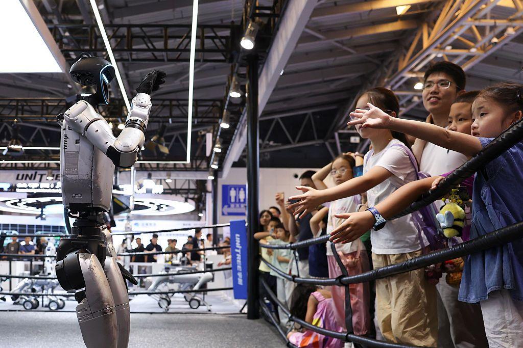 Un robot de Unitree, en un cuadrilátero, saluda a un grupo de niños en un evento con el público.
