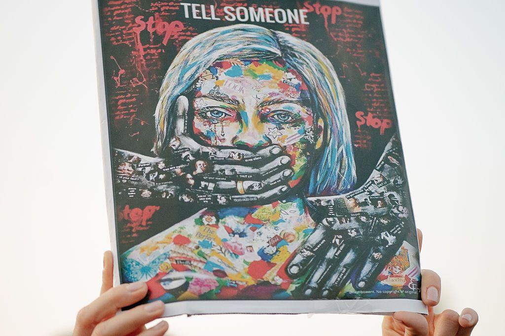 一幅示威的標語，圖畫中的女子被雙手掩著嘴巴，印有「Tell Someone」的字眼。