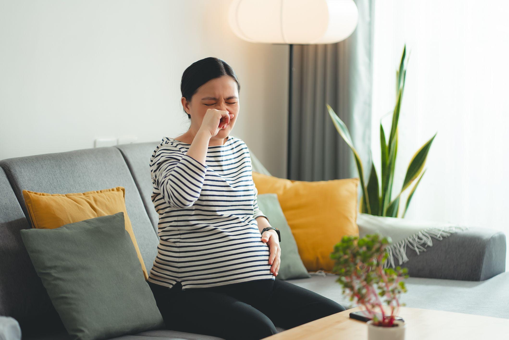 امرأة حامل ويبدو أنها متعبة، تضع يدها على أنفها، وتجلس على أريكة في غرفة المعيشة. وترتدي قميصاً بأكمام طويلة ومخطط باللونين الأبيض والأسود، وبنطال قماش أسود.