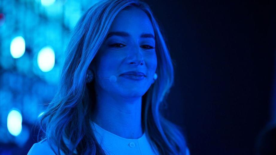 Mujer de 30 años, cabello largo rubio, atractiva; toda ella y el fondo se ven en tonos azules por la luz del lugar donde está