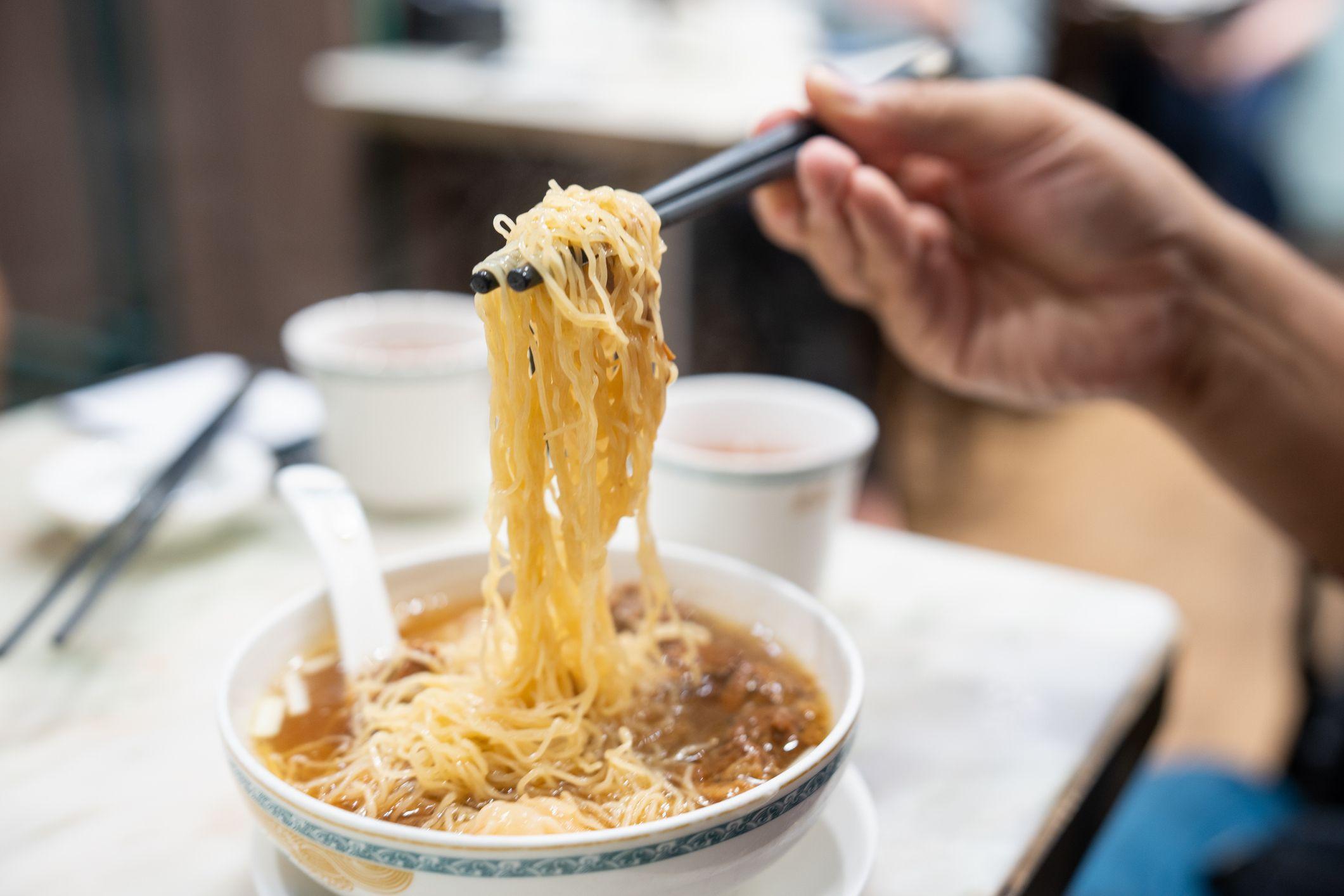 يد حنطية اللون لرجل يتناول طبقاً من نودلز باستخدام عيدان تناول الطعام في مطعم. الطاولة لونها أبيض والطبق الدائري كذلك لونه أبيض ومزركش بالأزرق ويصلح لتناول الحساء.