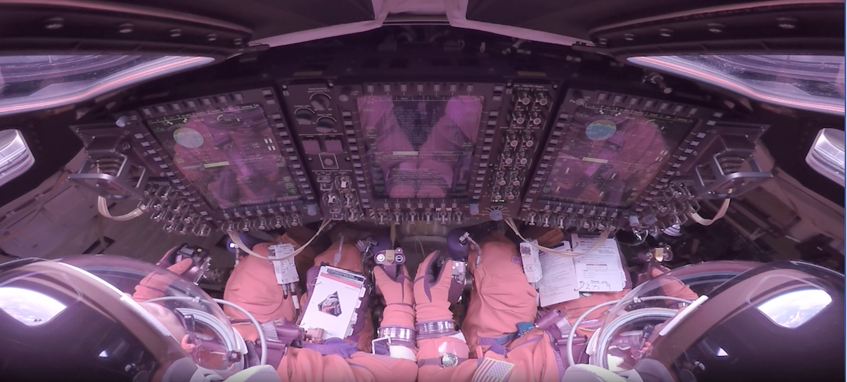 這張影像從兩名阿提米絲二號太空人的肩後視角向前拍攝，他們身穿橘色加壓太空衣，被牢牢固定在獵戶座乘員艙的座椅上。兩人戴著透明頭盔罩，面向一整面高聳、發光的顯示螢幕，以及密集排列的開關與旋鈕。戴著手套的雙手放在操控桿與檢查清單旁。畫面邊緣可見弧形窗戶、纜線與設備，使整個艙室顯得緊湊而高度技術化。