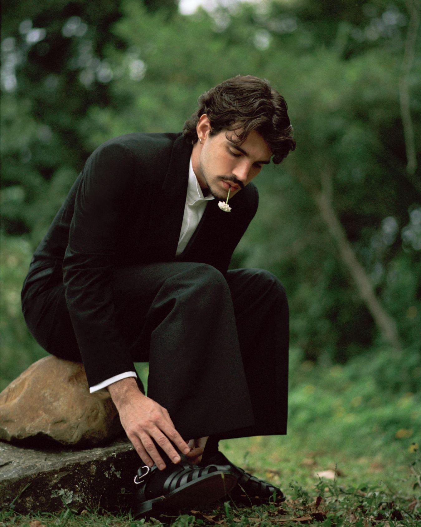 Jão, um homem de terno preto e camisa branca, está sentado sobre uma pedra em uma área gramada, inclinando-se para amarrar o sapato. Ele segura uma pequena flor branca entre os lábios, enquanto o fundo desfocado revela árvores e vegetação.