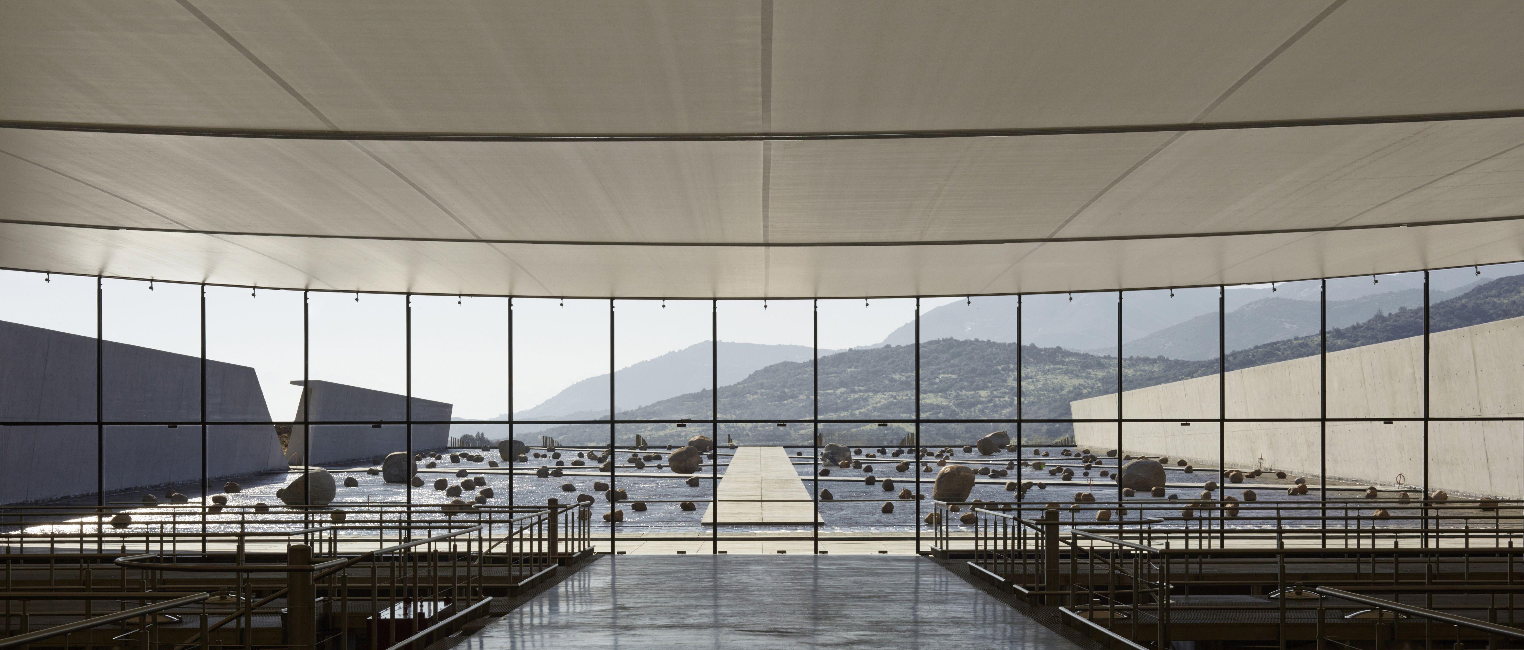 Vista interior hacia un gran ventanal que enmarca un patio con rocas, con montañas al fondo.