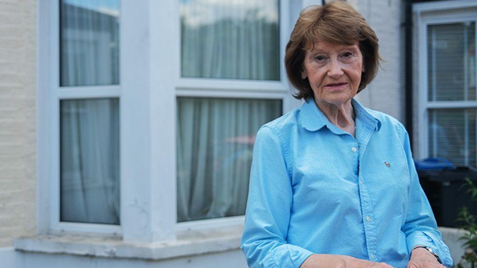 Sue está em frente a uma casa geminada de tijolos ou pedras de cor clara. Ela está vestindo uma camisa azul clara de botões com mangas enroladas até os cotovelos e um pequeno logotipo bordado no lado esquerdo do peito. Atrás dela, a casa tem duas grandes janelas brancas com cortinas transparentes e uma saliência abaixo de cada janela. A cena geral é bem iluminada e não há objetos ou texturas adicionais visíveis