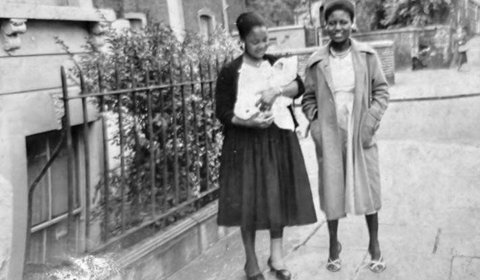 Ijeoma en brazos de su madrina y su madre al lado sonriendo en una foto en blanco y negro. Las dos mujeres adultas van vestidas con abrigo largo, por debajo de las rodillas y vestidos a la misma altura. Están en la calle, junto a la reja de una casa.
