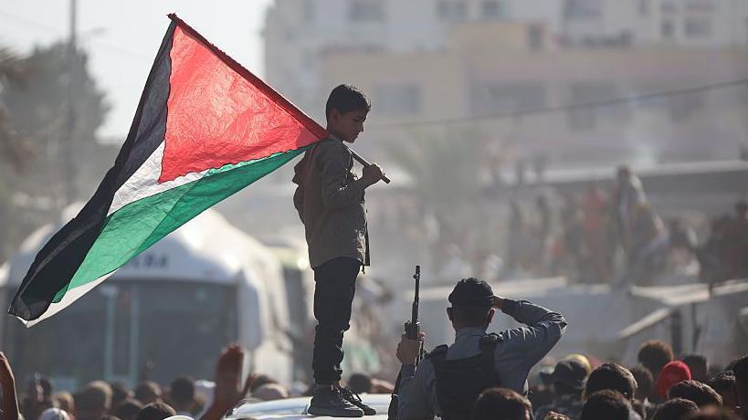 Autobuses que transportan a palestinos liberados de prisiones israelíes en virtud de un acuerdo de alto el fuego en Gaza y de intercambio de rehenes con facciones palestinas llegan al hospital Nasser en Jan Yunis, Franja de Gaza. En primer plano, un niño subido a una plataforma con una bandera de Palestina.