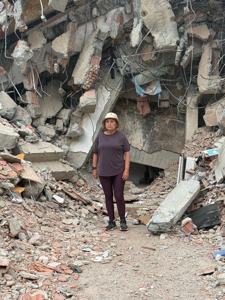 María Lourdes Pérez, vestida con un pantalón y camisa marrones y un gorro beige. Detrás de ella hay una montaña de escombros, hormigón, piedras, partes de una edificación