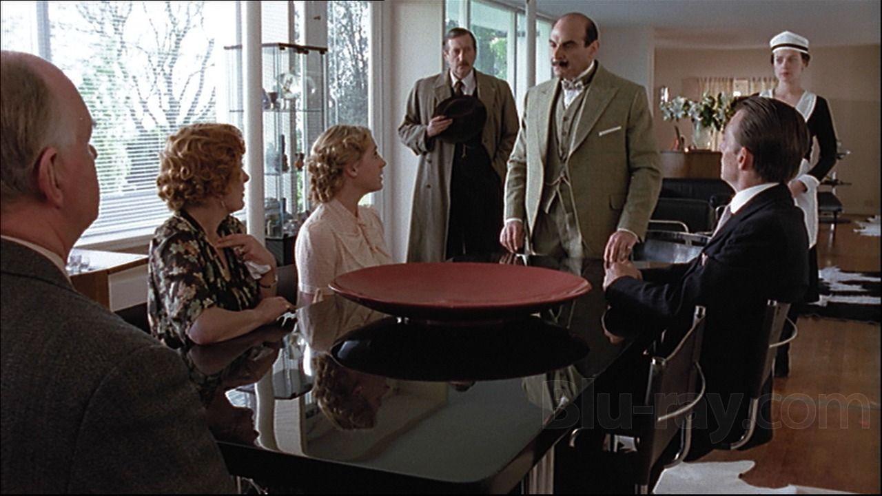 Cena em ambiente interno mostra seis pessoas reunidas ao redor de uma mesa preta com um objeto circular vermelho no centro. Um homem de terno claro est&aacute; em p&eacute; falando com os demais, enquanto outro homem aparece ao fundo segurando um chap&eacute;u. &Agrave; direita, uma mulher vestida como empregada dom&eacute;stica observa a cena.