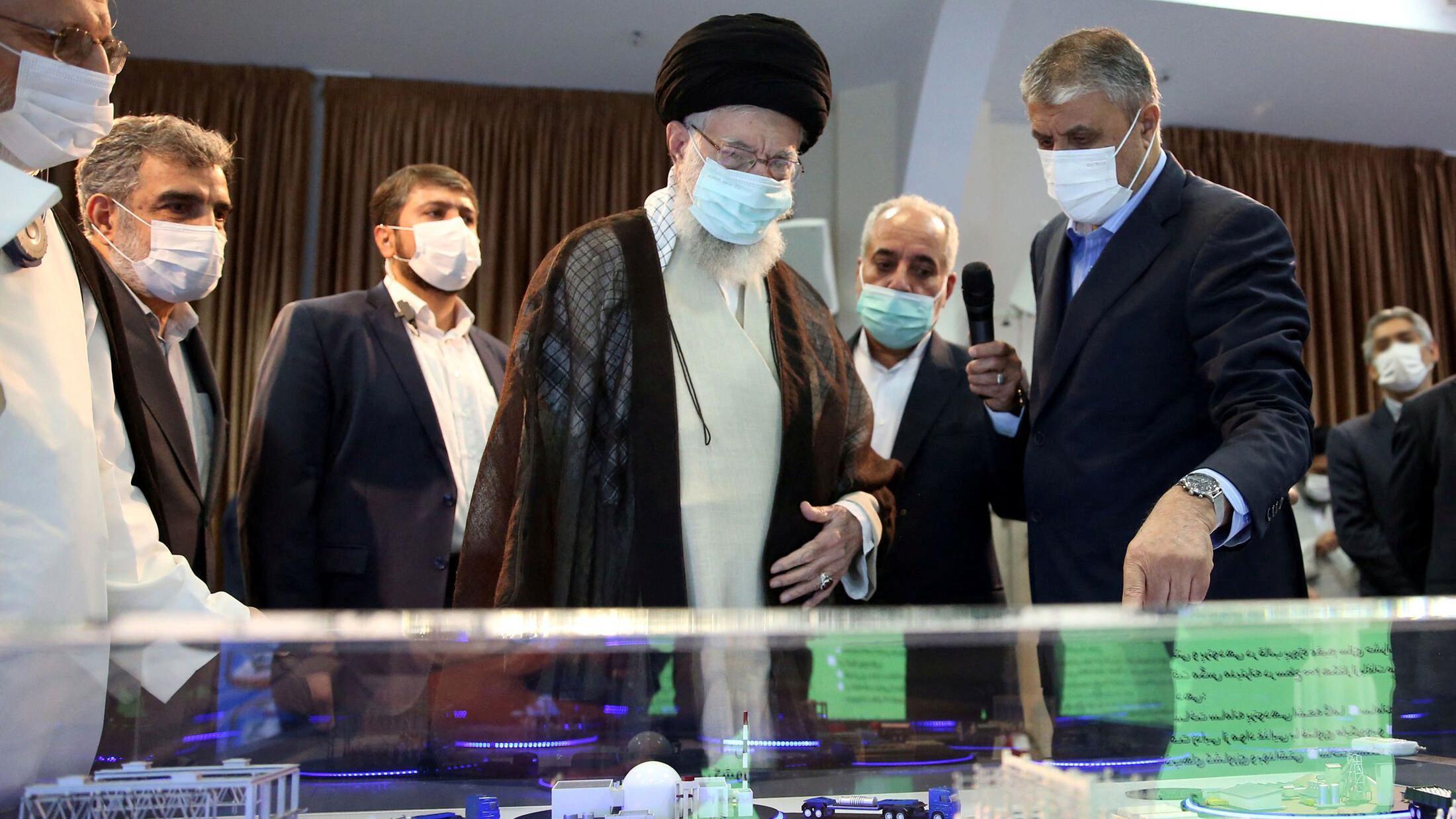 Khamenei views a model of a nuclear facility, in Tehran on 11 June 2023. He is wearing a blue face mask and traditional clothes while other men stand around him. One is wearing a white laboratory jacket. The model is in a display cabinet and consists of a white reactor dome and other buildings in one location, along with the framework of a rectangular building with models of people in it nearby. 