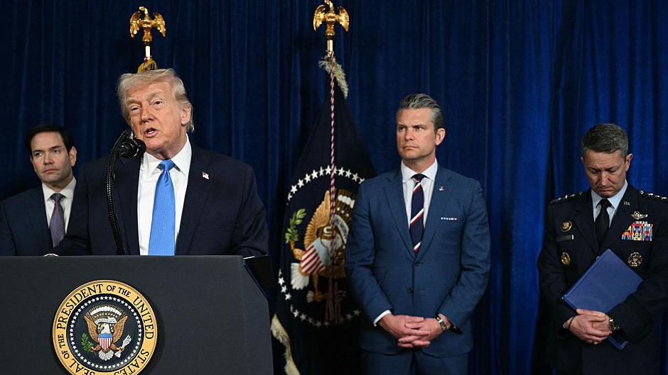 Donald Trump se encuentra en el podio dirigiéndose al público, con una corbata azul y gesticulando con una mano. Detrás de él se encuentran el Secretario de Estado Marco Rubio, el Secretario de Defensa Pete Hegseth y el Presidente del Estado Mayor Conjunto de Estados Unidos, el General Dan Caine, vestido con uniforme militar.