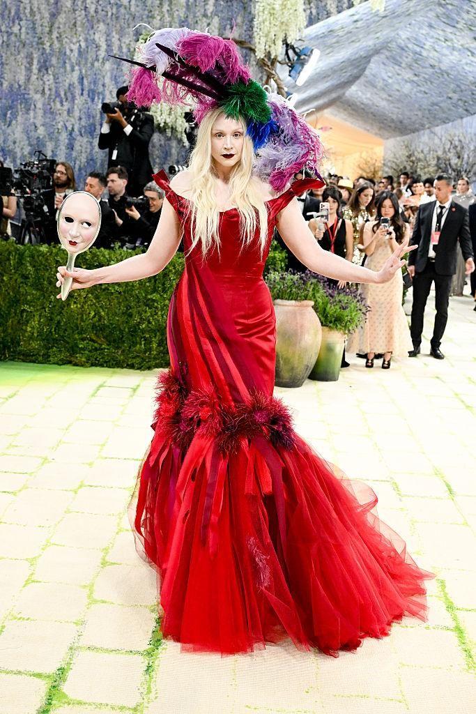 Una mujer posa en una alfombra verde durante un evento de gala, con un vestido rojo voluminoso, un tocado de plumas multicolor y una máscara en la mano, rodeada de fotógrafos y asistentes.