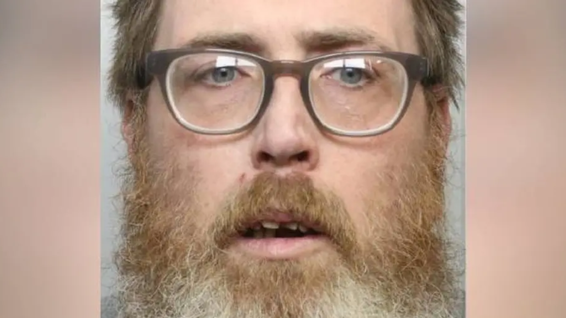 Mugshot photograph of Dale Morgan, 42. He has a long shaggy ginger and grey beard, short ash strawberry blonde hair and blue eyes. He wears square rimmed reading glasses and looks confused into the camera, with his mouth part open revealing some of his teeth. 