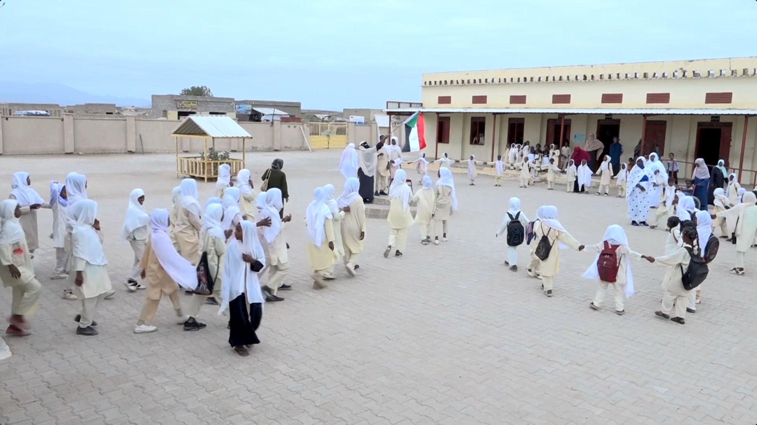 طلاب يرتدون الملابس الرسمية ويحملون حقائبهم، ويشكّلون مجموعات في ساحة المدرسة مع انطلاق الدراسة فيها، وتقف بينهم معلماتهم.