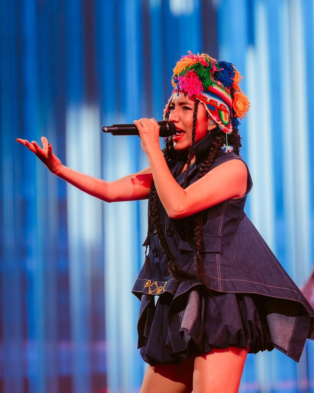 Renata Flores en el escenario, lleva un vestido de corte muy moderno de tela de jean y lleva un chullo, un gorro de lana tradicional peruano de muchos colores. 