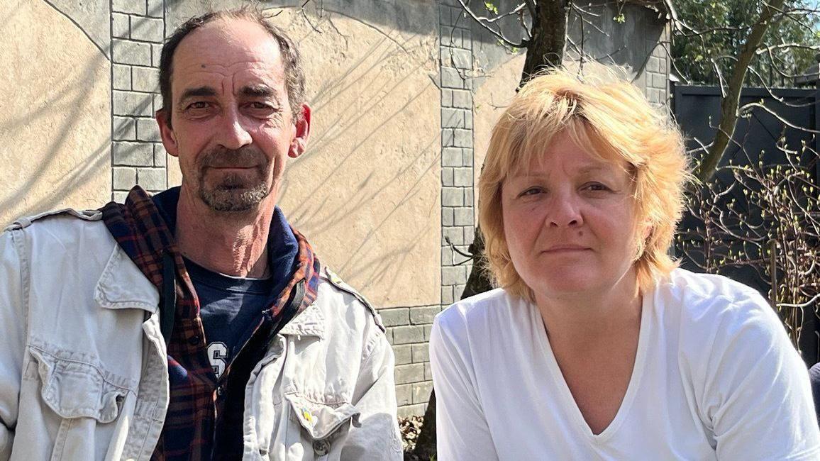 Un hombre y una mujer de mediana edad, sentados frente a una pared y un árbol, mirando a la cámara.