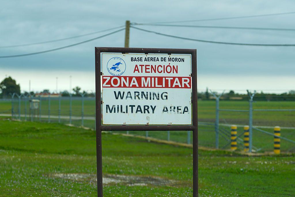 Cartel donde se lee "Base Aérea de Morón. Atención, zona militar. Warning Military Area". Al fondo se ve una llanura verde cercada con vallas. 
