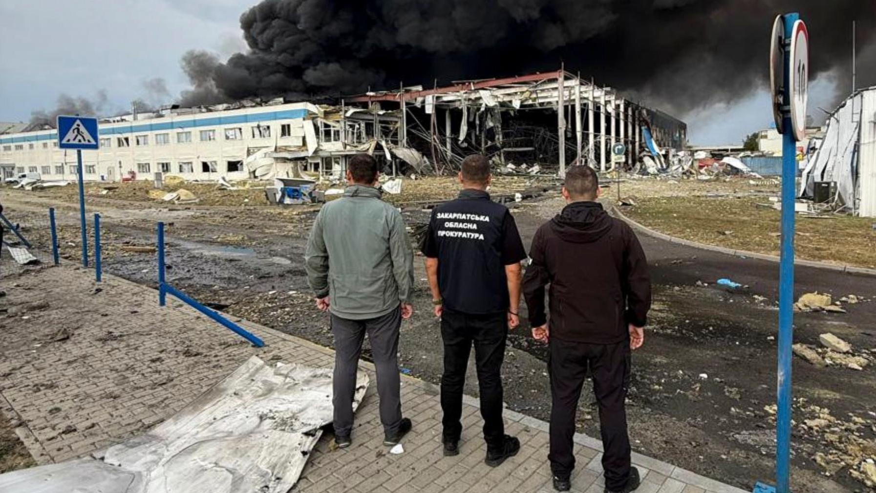 Tres hombres están de espaldas a la cámara mientras miran los escombros de un edificio que aún se quema y lanza una enorme columna de humo negro al cielo
