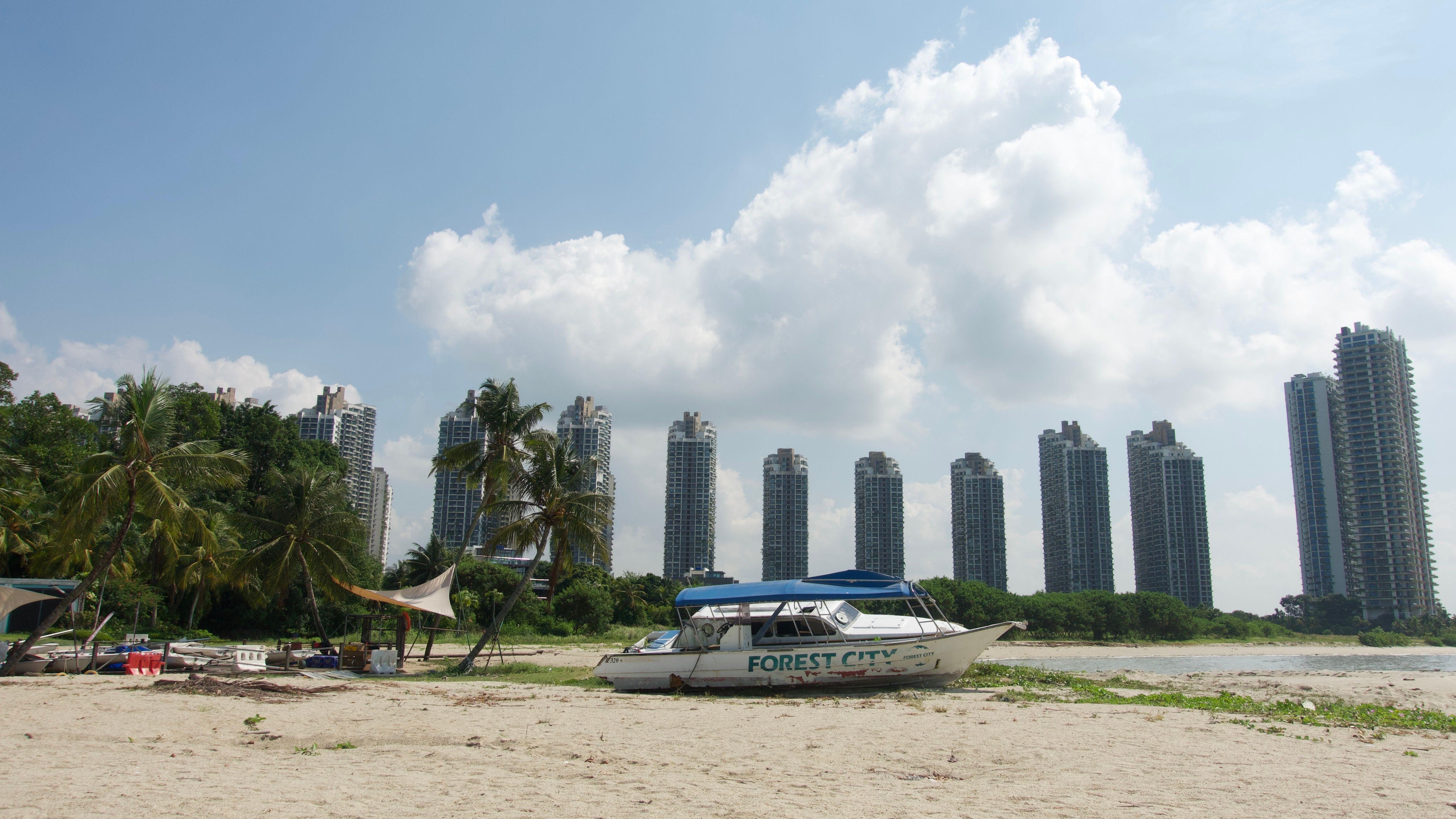 Sebuah kapal tua yang rusak bertuliskan 'Forest City' di sisinya berada di atas pasir pantai, di samping tumpukan puing acak dan pohon palem, dengan gedung-gedung bertingkat tinggi di latar belakang yang jauh.