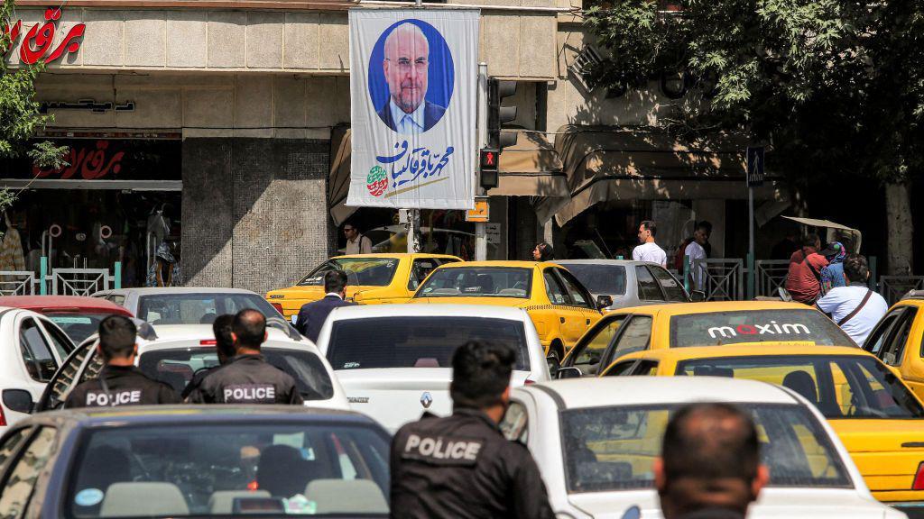 A photo of Mohammad Bagher Ghalibaf on a flag, hanging above congested roads of yellow and white cars.