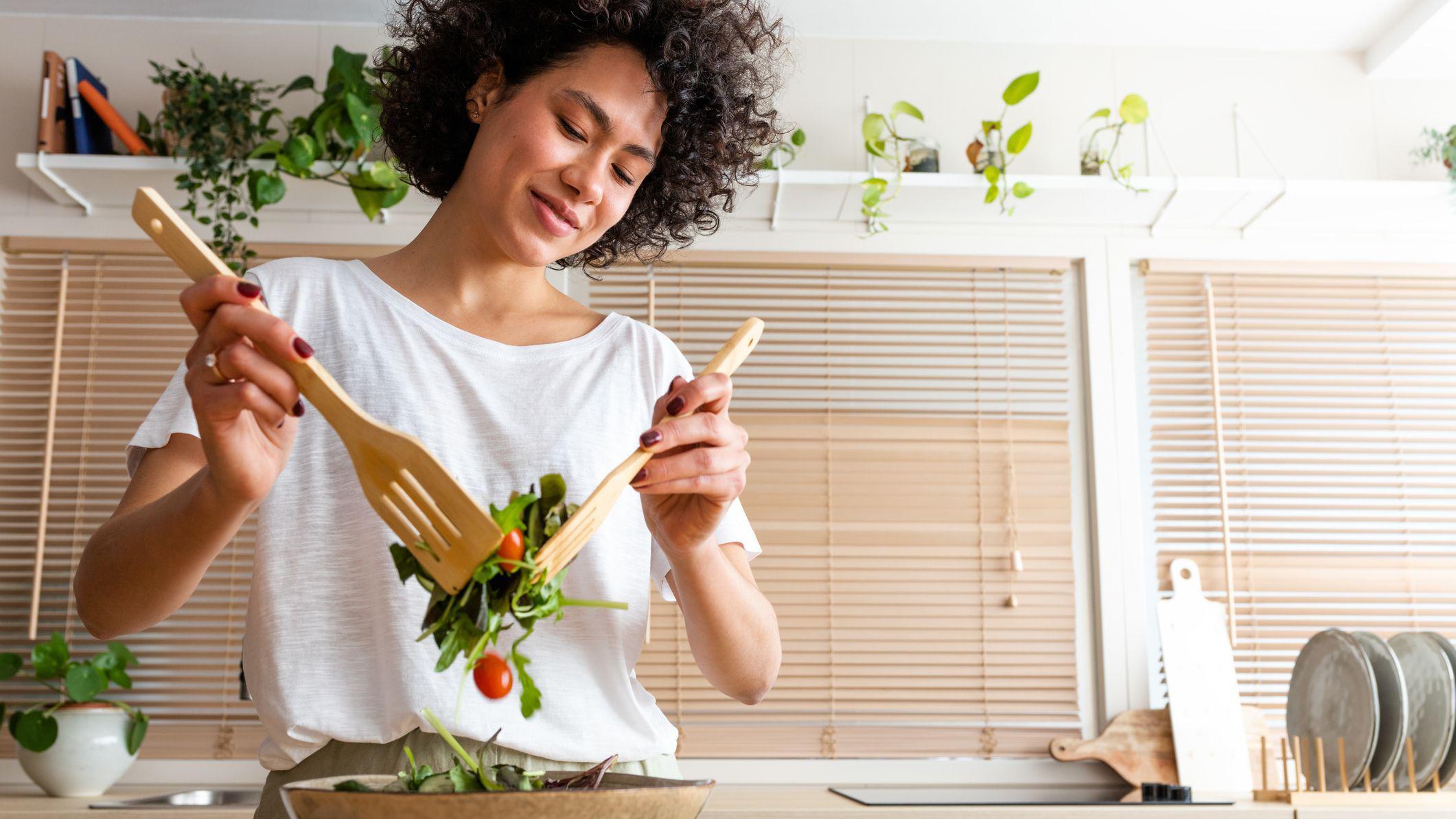 Mulher preparando salada em uma cozinha moderna, usando utensílios de madeira para misturar verduras e tomates em uma grande tigela