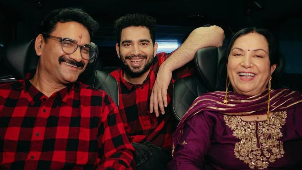 Samay Raina poses gleefully with his mum and dad inside a car. He and his father are wearing matching red-black check shirts, while his mum is wearing a purple tunic and scarf with golden threadwork. 