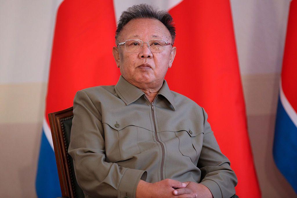朝鮮前領導人金正日身穿墨綠色襯衫，戴著眼鏡，坐在椅子上，背景是朝鮮國旗