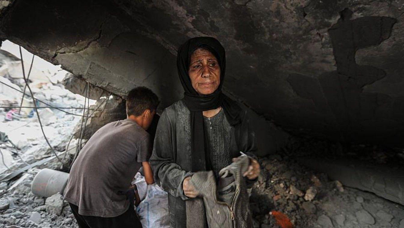 امرأة فلسطينية ترتدي حجاباً داكناً وفستاناً رمادياً، وهي تحمل قطعة ملابس في يدها، تنظر بحزن، بينما تتفقد ما تبقى من الأنقاض في سوق غزة الذي تعرض للقصف الإسرائيلي. ويقف خلفها صبي متجهاً بظهره إلى الكاميرا ويرتدي قميصاً رمادياً قصير الأكمام وينطالاً أسود.