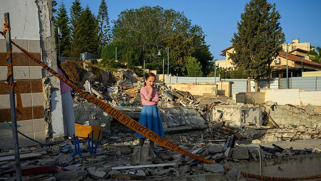 Una niña reacciona ante las secuelas de un ataque con misiles iraní, de pie entre los escombros junto a lo que fue la sinagoga de Beit Shemesh, Israel, el 5 de abril de 2026. Con una falda azul y una camiseta rosa, y el pelo recogido en una coleta, lleva un brazo cruzado sobre el pecho mientras que con el otro se tapa el rostro, visiblemente conmocionada. La zona del ataque está acordonada con cinta amarilla con inscripciones en hebreo. 