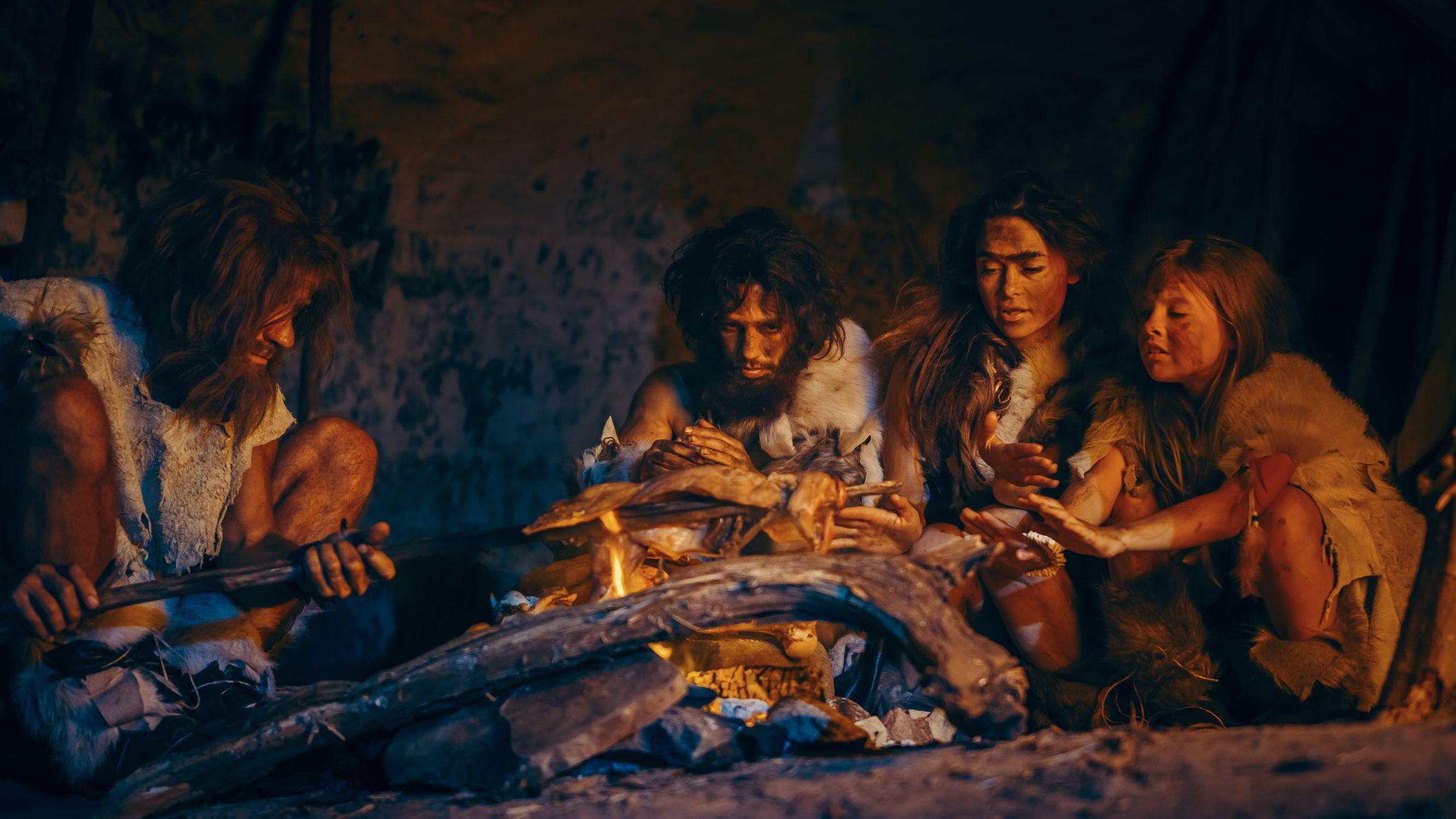 Tribu de cazadores-recolectores prehistóricos vestidos con pieles de animales asando y comiendo carne en una cueva por la noche.
