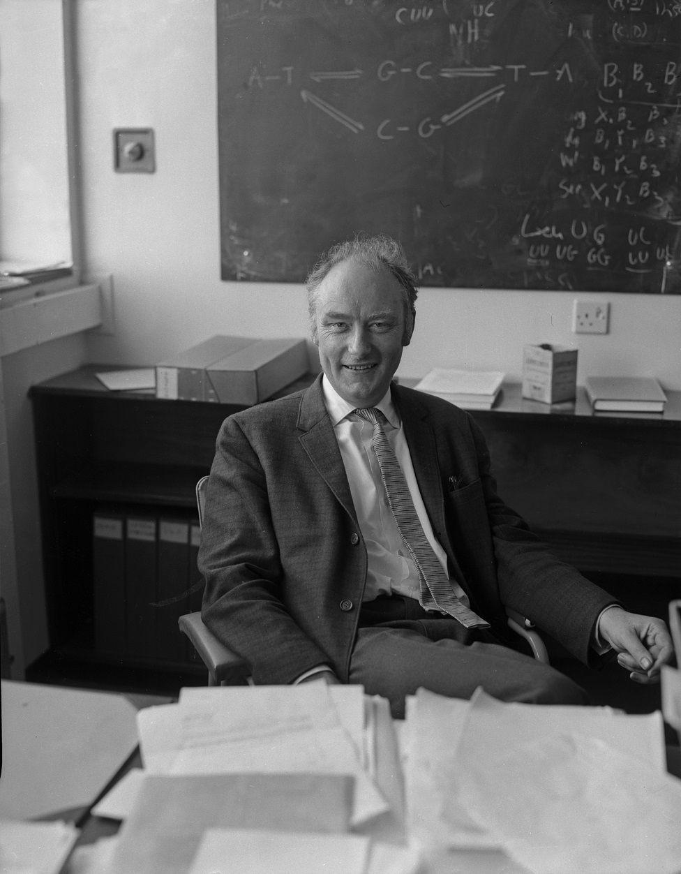 Francis Crick sentado en un escritorio con un pizarrón detrás