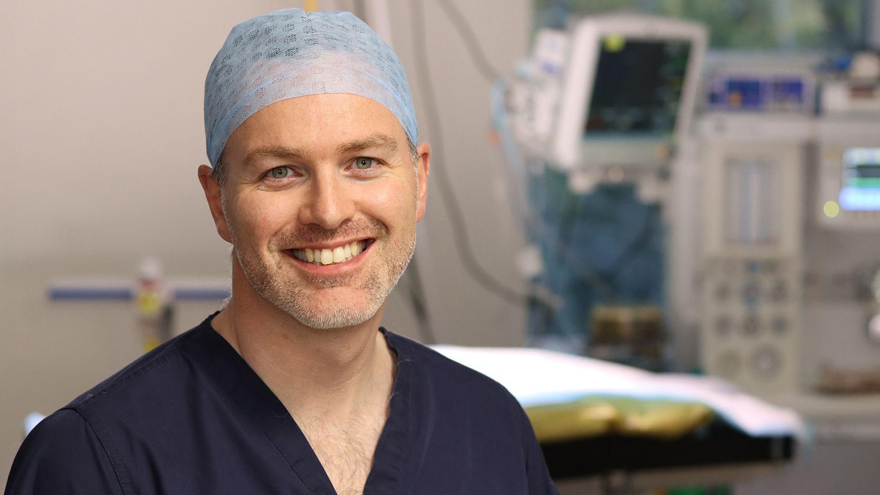 一名男子在手術室中微笑，頭戴髮網並穿著深藍色醫療手術服