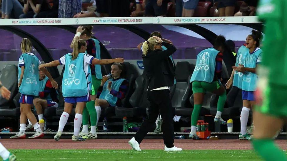 Wiegman, parada frente a las bancas con las manos en la cabeza, mientras las jugadoras suplentes calientan.