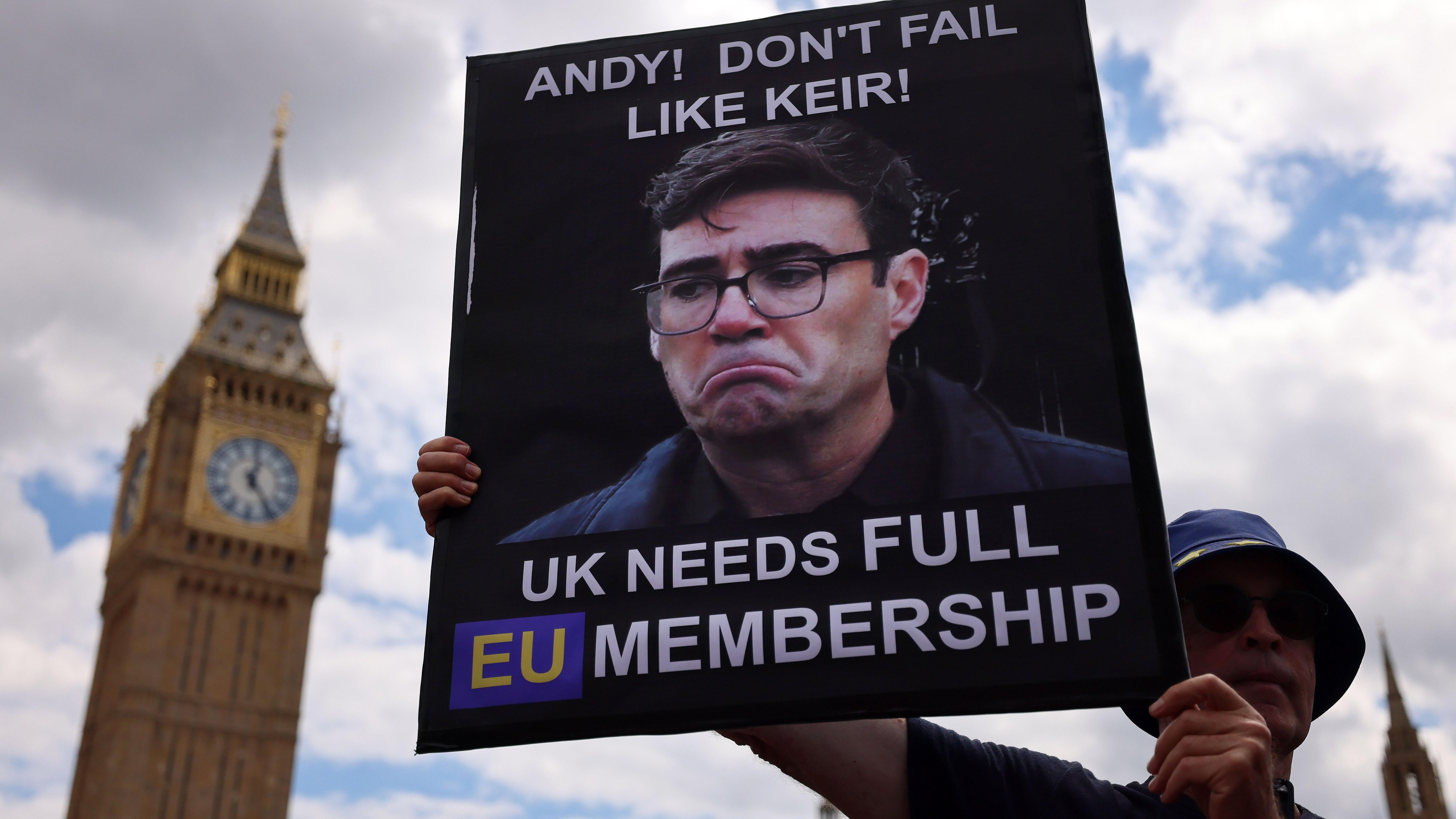 Um ativista pr&oacute;-Uni&atilde;o Europeia, usando um chap&eacute;u, est&aacute; do lado de fora do Parlamento Brit&acirc;nico segurando um cartaz com a imagem de Andy Burnham com uma cara chateada. O cartaz diz: 