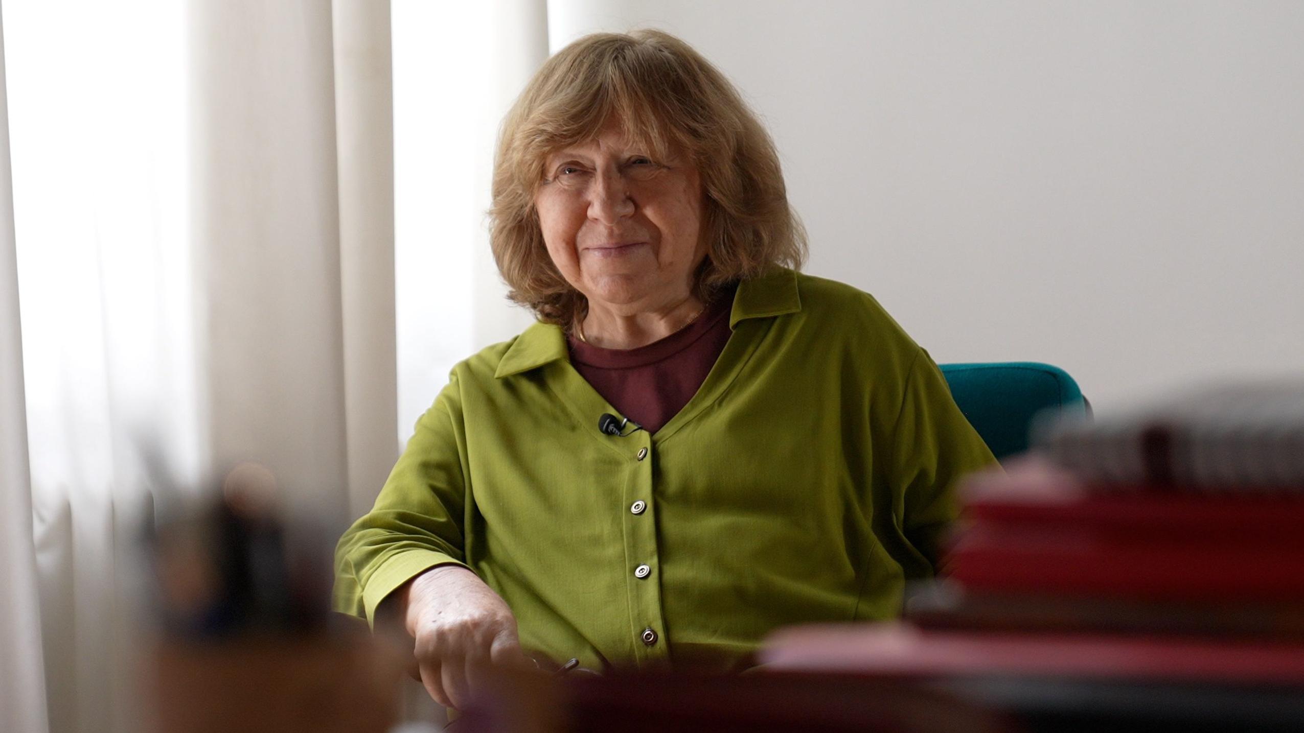 Una sonriente Svetlana Alexievich está sentada detrás de una pila de libros en su mesa de trabajo, vistiendo una blusa verde.