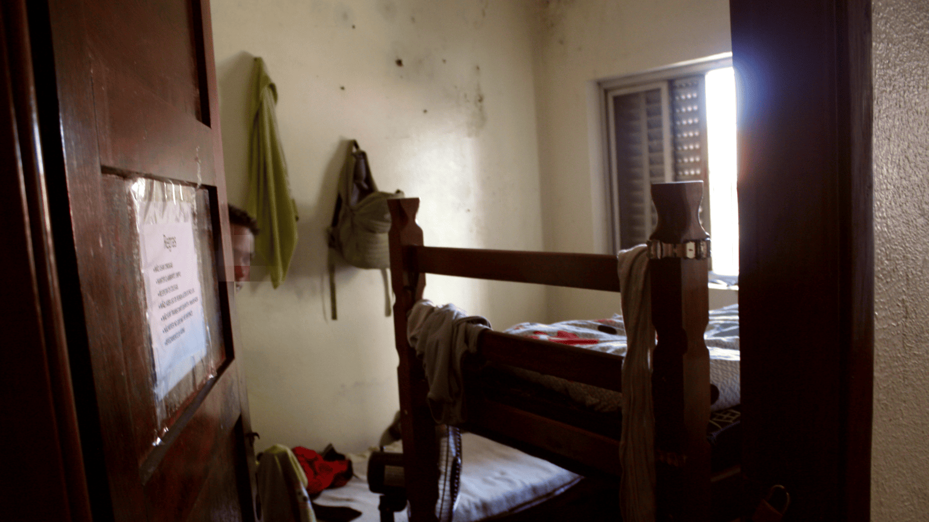 Pessoa, com rosto ocultado, abre porta de quarto, onde se pode ver beliche, paredes manchadas e roupas penduradas em diversos pontos